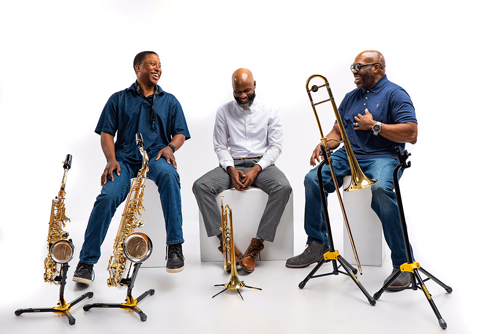 3 members of mo'horns posing with their instruments on stands